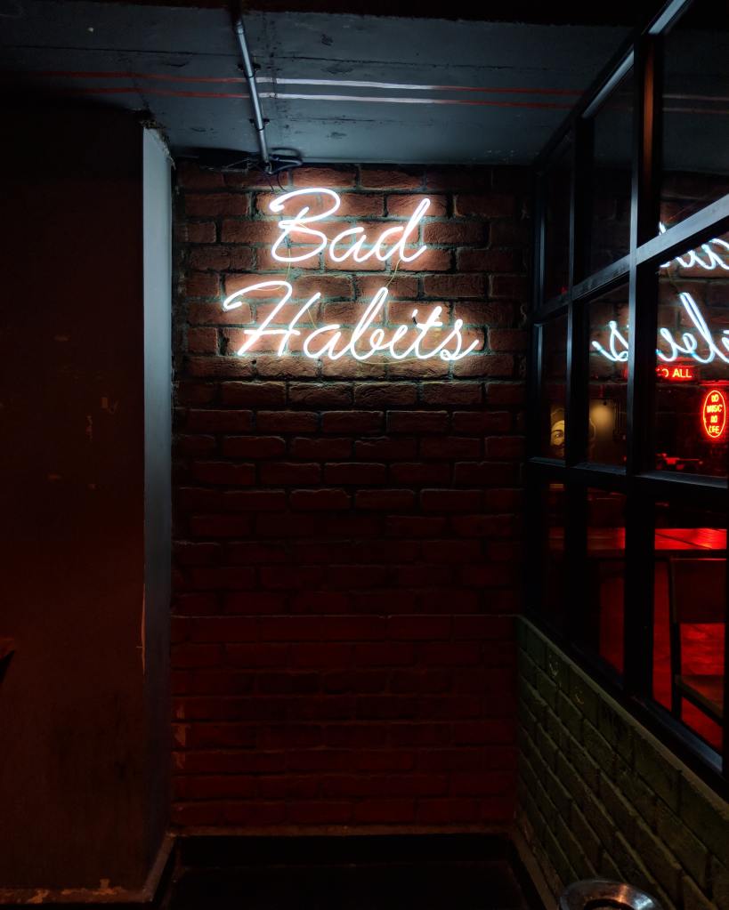 White neon sign reading 'Bad Habits', mounted on a brick wall.