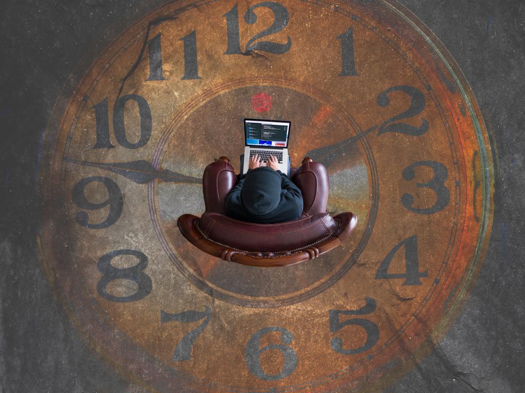 Person in a chair with a laptop, sitting at the centre of a clockface. 